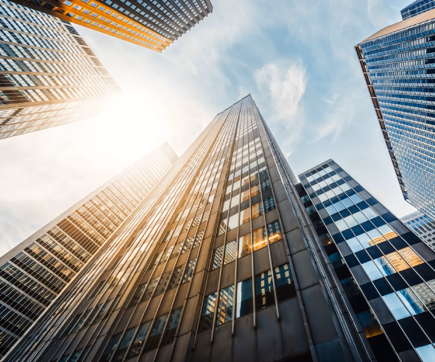 Looking up at tall modern office buildings with reflective glass under partly cloudy sky.