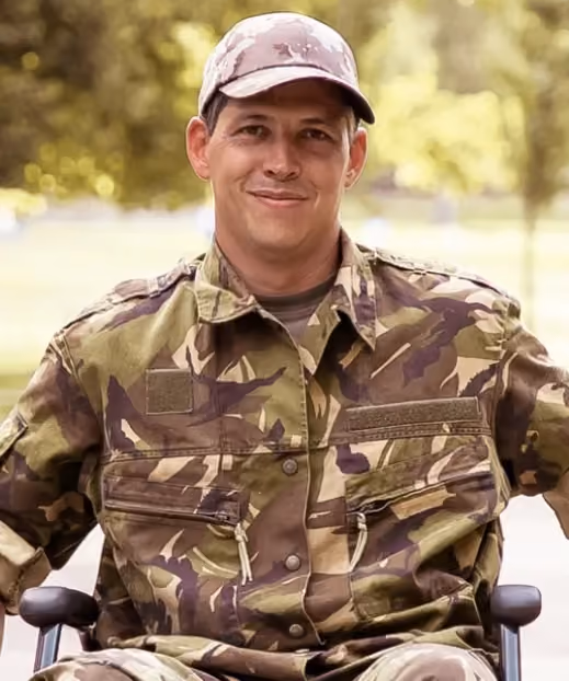Smiling soldier in camouflage uniform and cap sitting outdoors in a wheelchair.