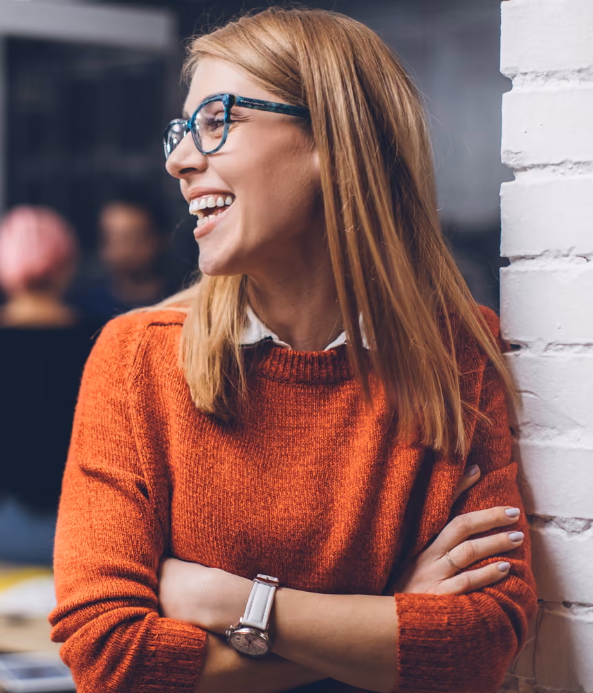 Smiling woman with glasses wearing a red sweater and watch leaning against a white brick wall.