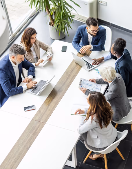Six businesspeople in formal attire having a meeting around a modern conference table with laptops and documents.