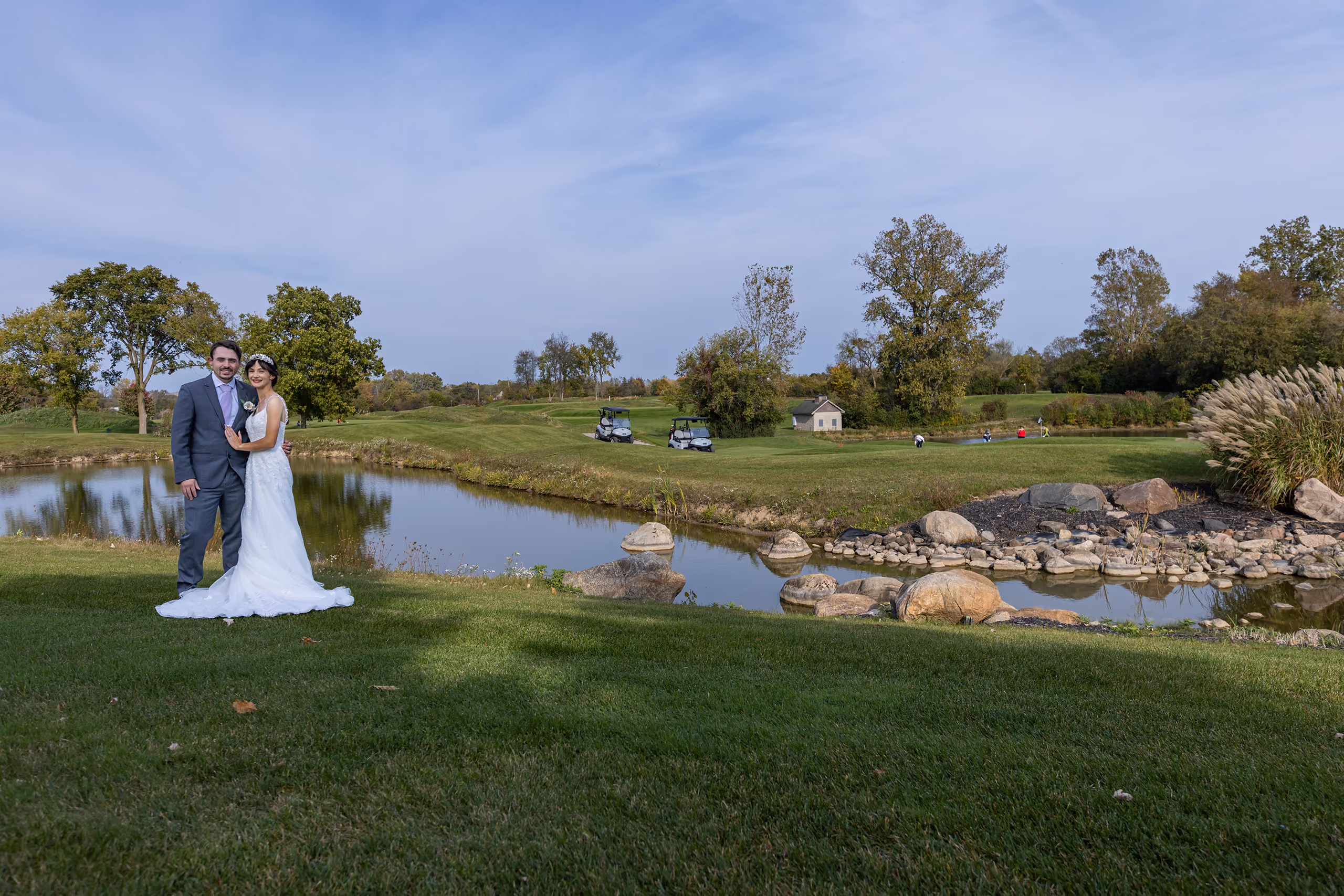 Wide shot of bride and groom standing by a golf course pond with rolling green fairways