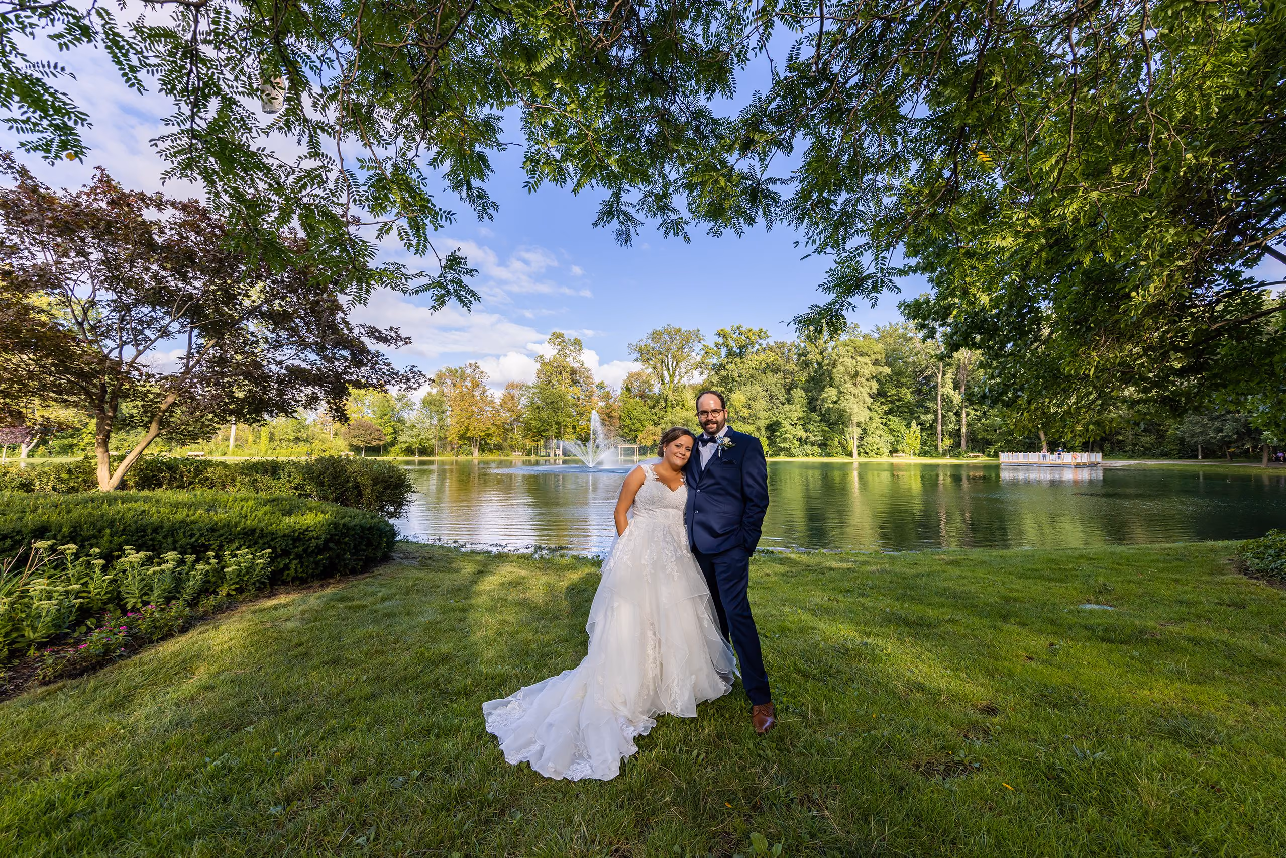 Bride and groom standing on green grass beside a lake with a fountain, Southeast Michigan wedding photography