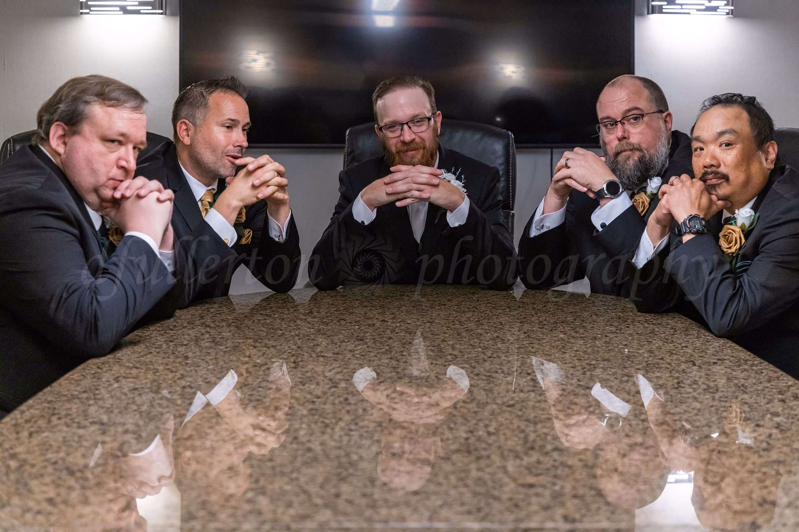 Groom and groomsmen posing seriously at a boardroom table, hands clasped like a business meeting