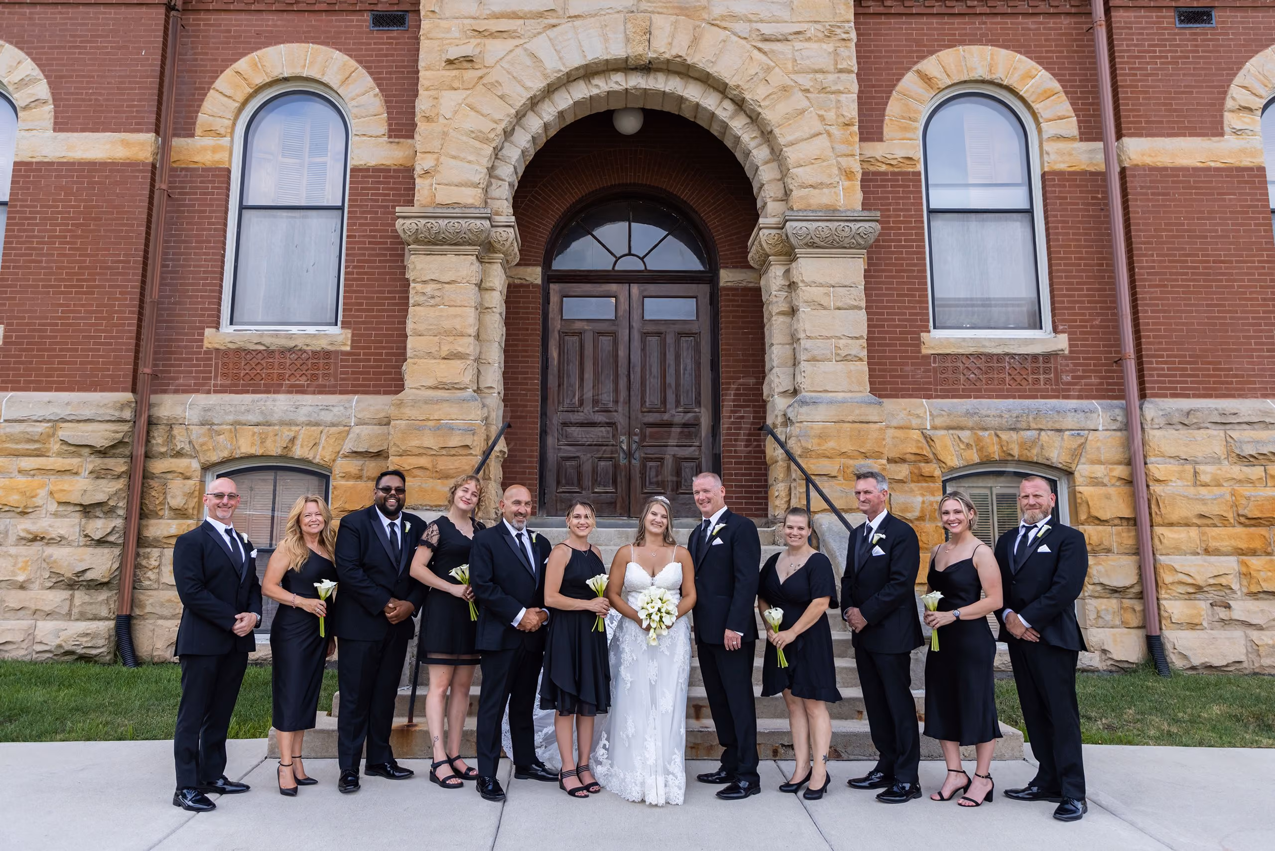 Wedding party posing under a grand stone archway of a historic brick building, bride and groom centered, Howell, MI