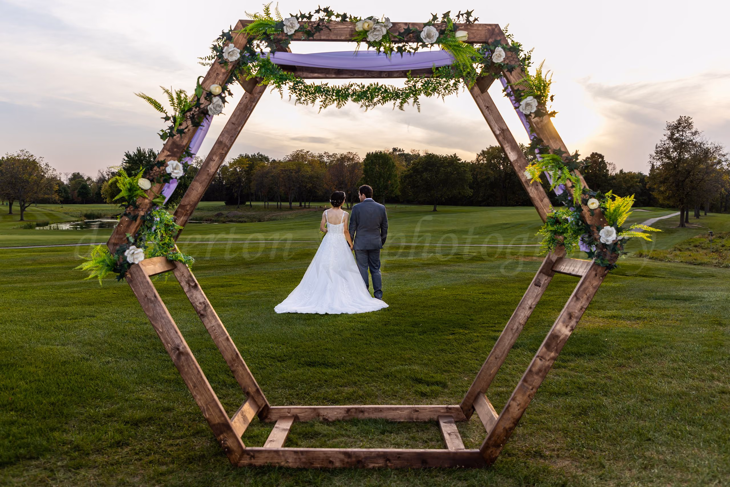 Bride and groom walking away hand in hand, framed by a large hexagonal floral wooden arbor at sunset