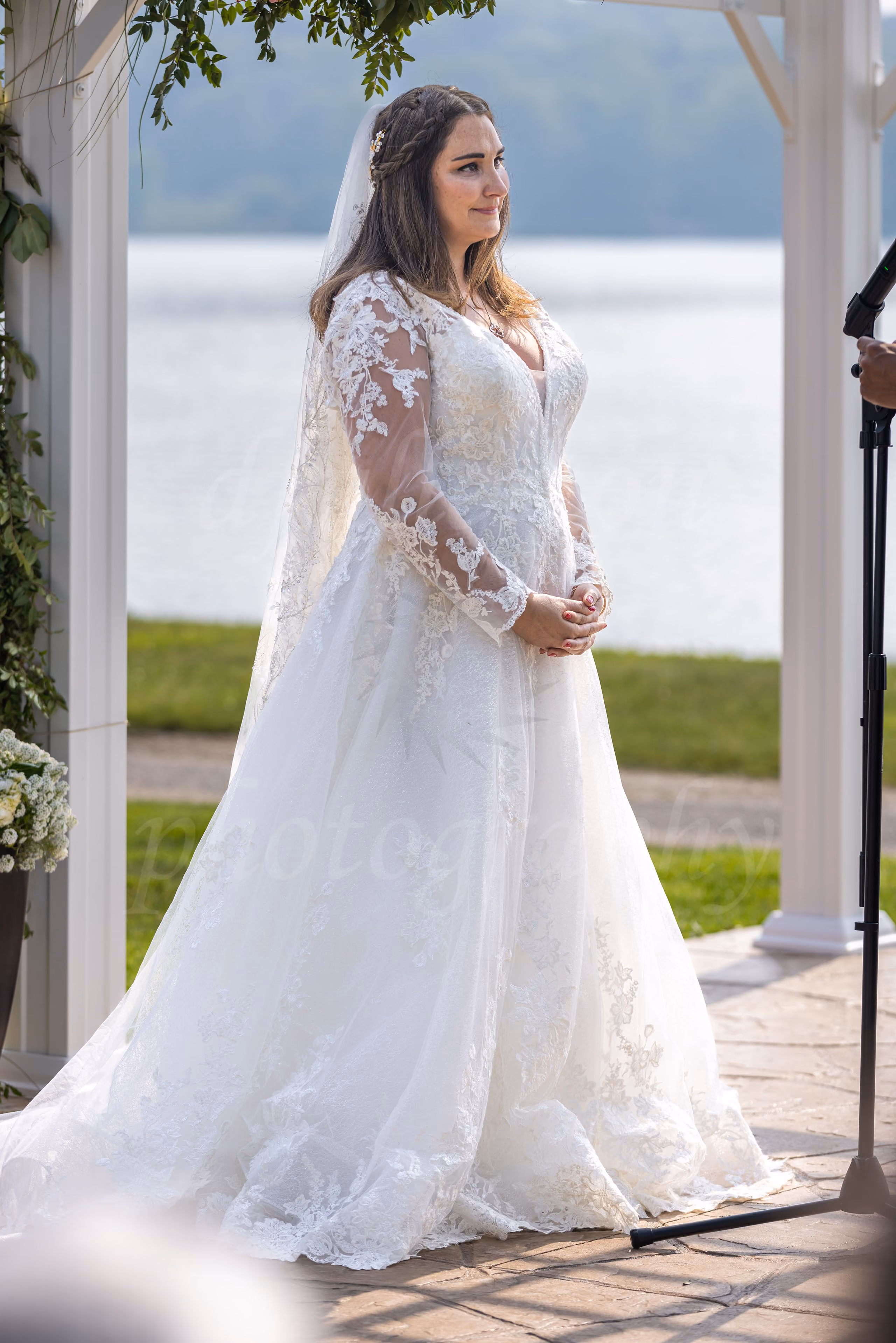 Bride in a long lace-sleeve gown and veil standing at the microphone during her outdoor lakeside ceremony
