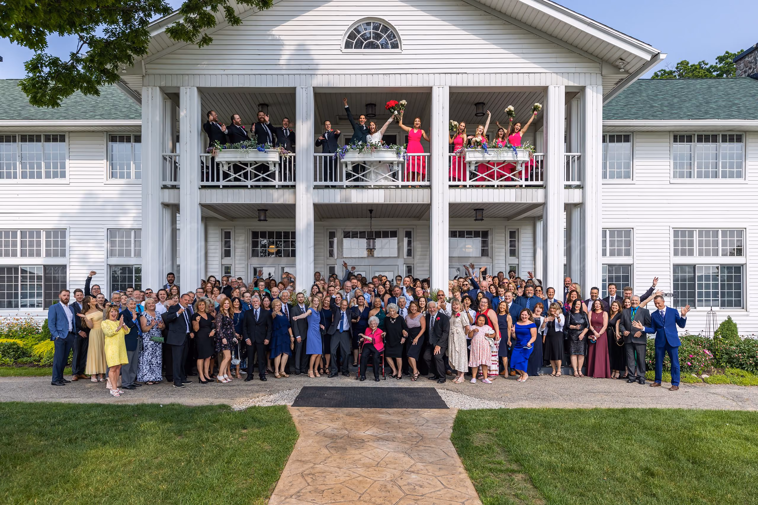 Bride and groom on a balcony with wedding party above a large crowd of guests gathered outside a white venue