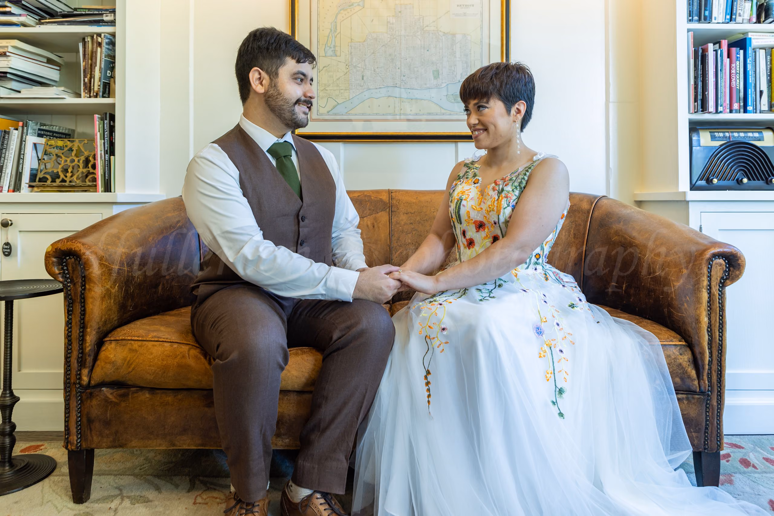 Bride and groom holding hands seated on a brown leather couch, bride in a floral-embroidered gown