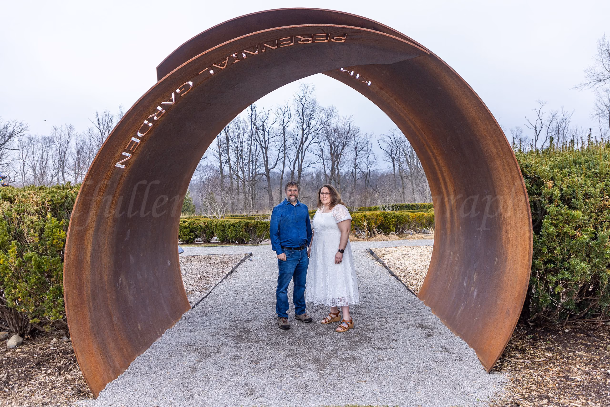 Couple standing under a large circular metal garden archway reading Perennial Garden