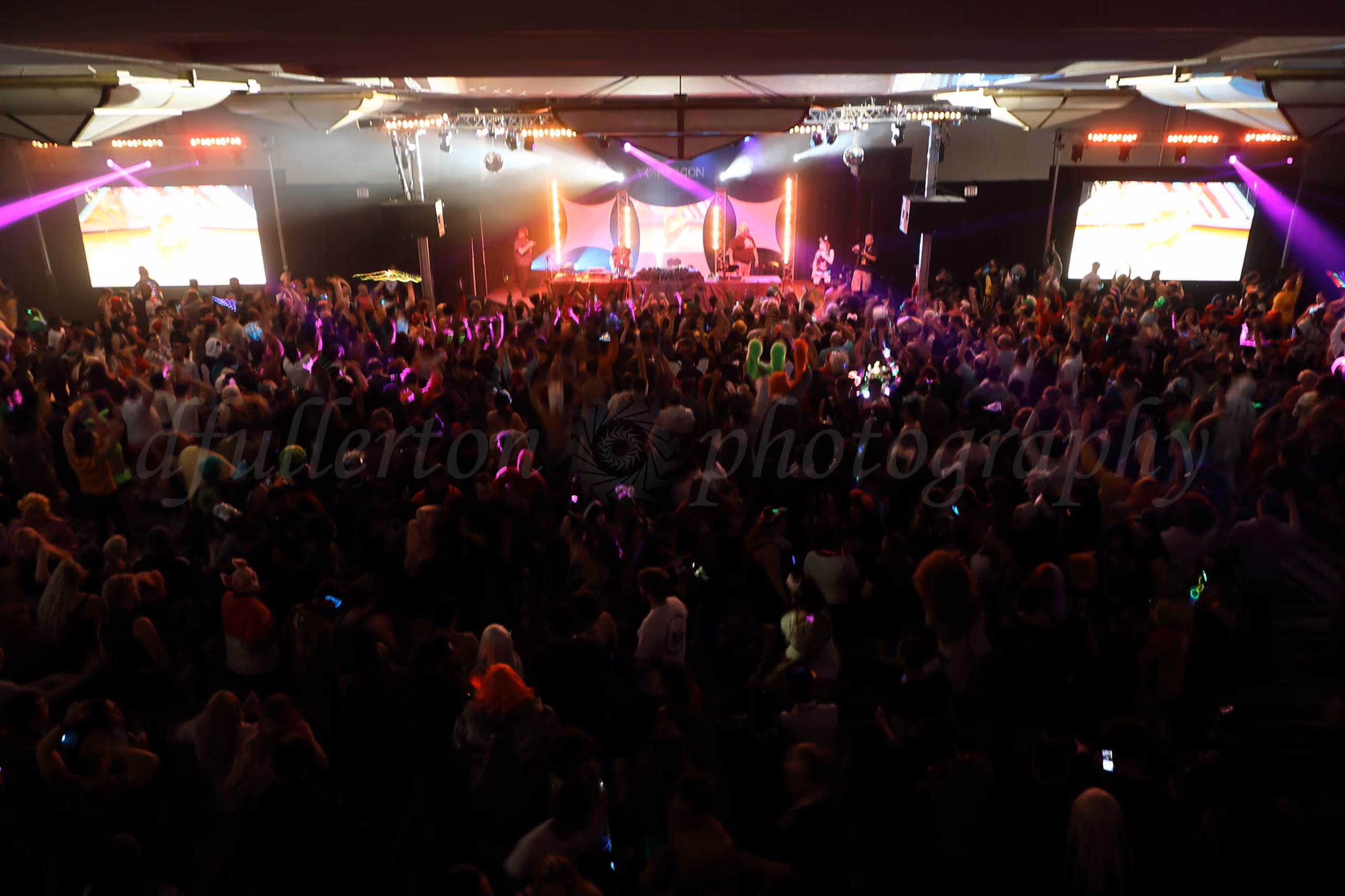 Large crowd dancing at a nighttime convention rave with colorful stage lights and video screens