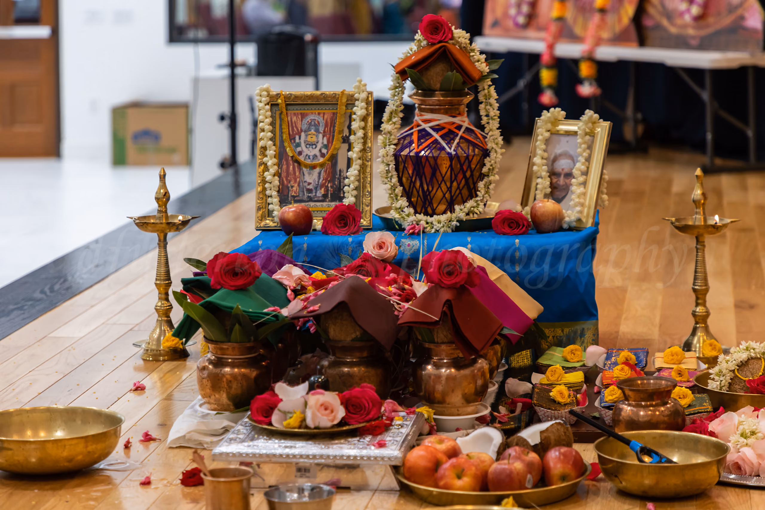 Traditional Hindu ceremony altar with brass kalash vessels, framed portraits, roses, and fruit offerings