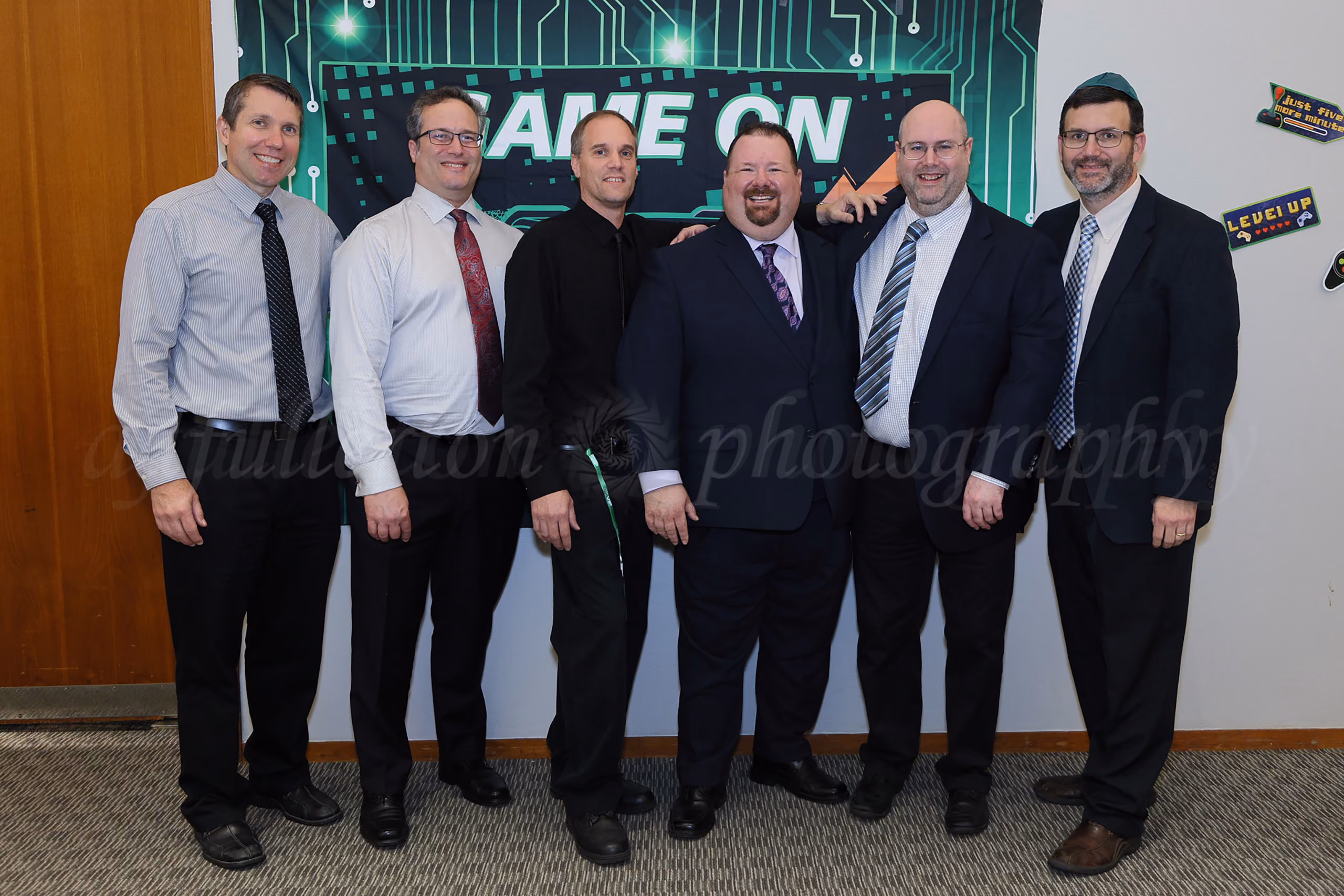Six men in business attire posing together in front of a Game On themed banner at an event