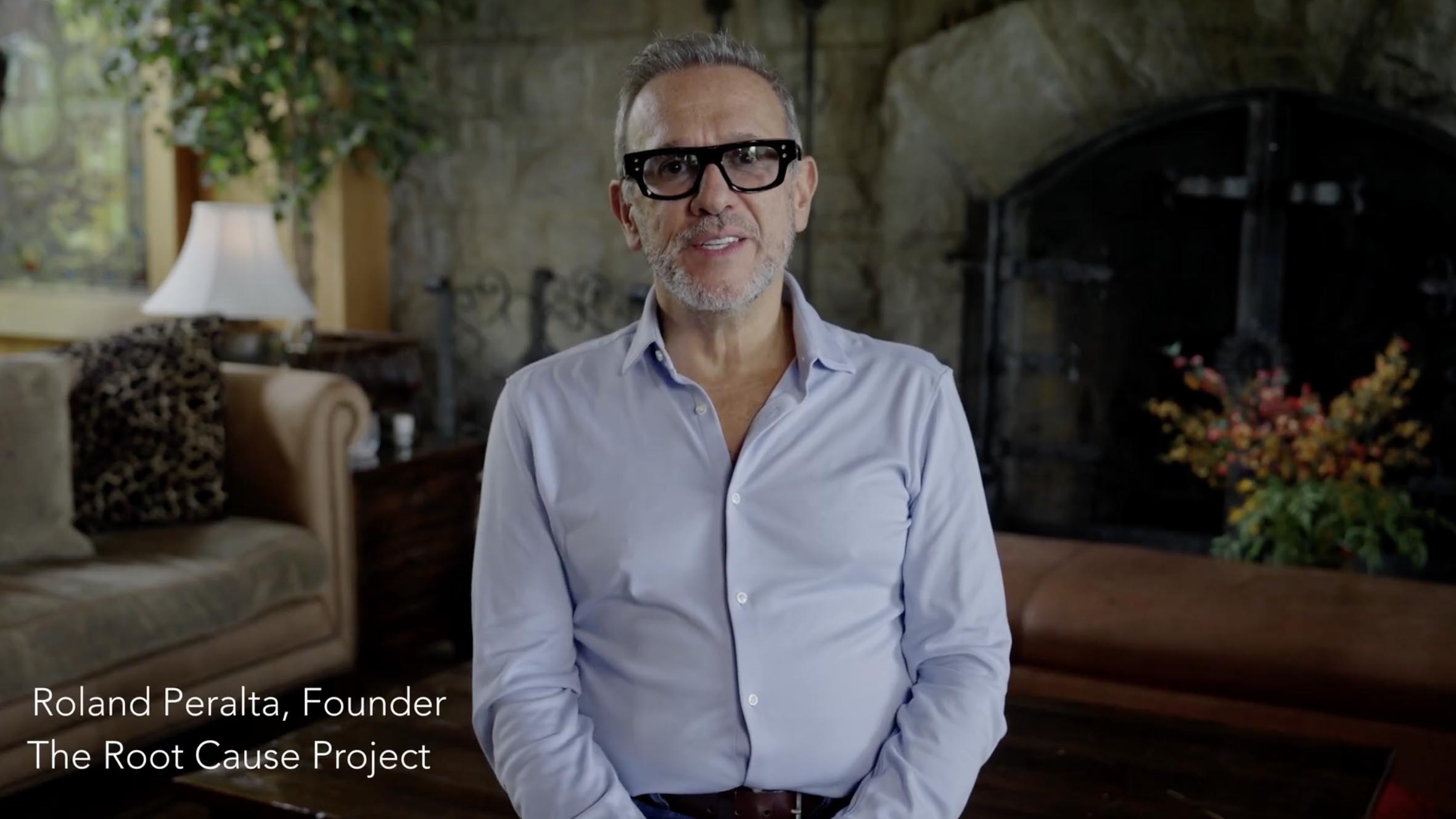 A man with short gray hair, a gray beard, black glasses, and a light blue button-up shirt is sitting in a cozy room with a stone fireplace, a brown leather couch, plants, and a lamp. Text on the image reads: Roland Peralta, Founder The Root Cause Project.