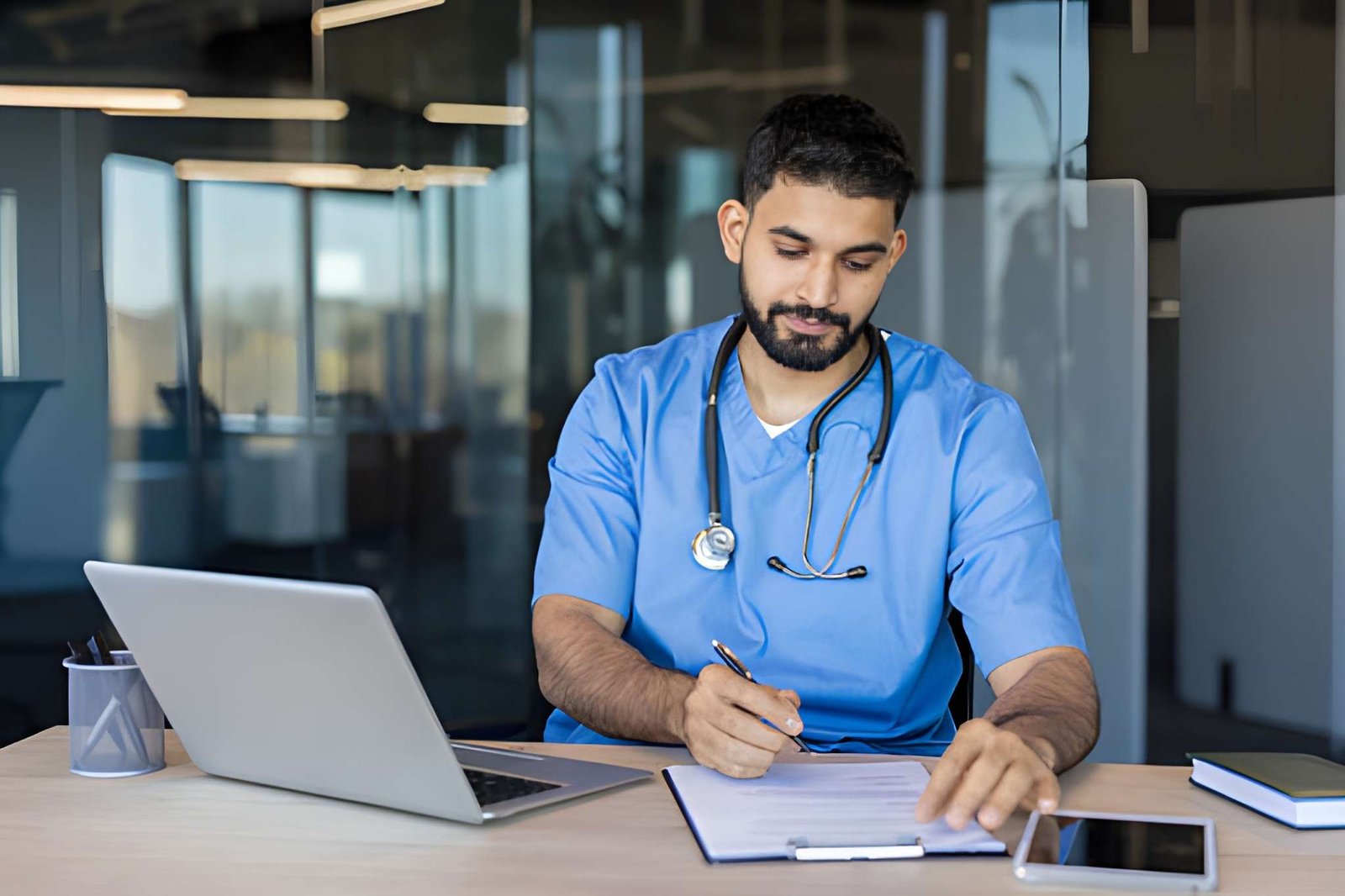 Male healthcare professional in blue scrubs reviewing documents at a desk with a laptop, representing clinical and medical mental health professionals.