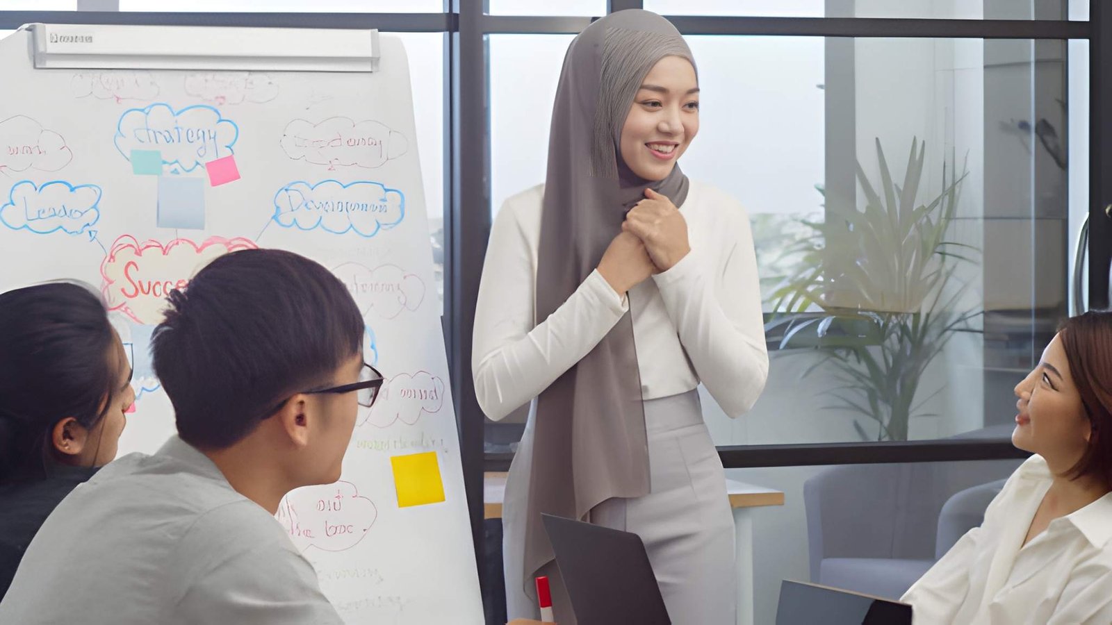 A smiling woman wearing a grey hijab leading a group presentation or coaching session.