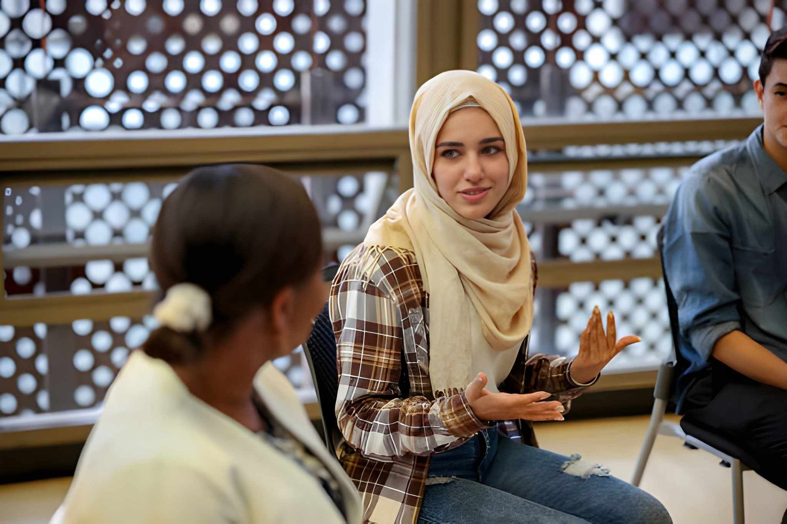 A young woman wearing a cream hijab gesturing while speaking in a group support setting.