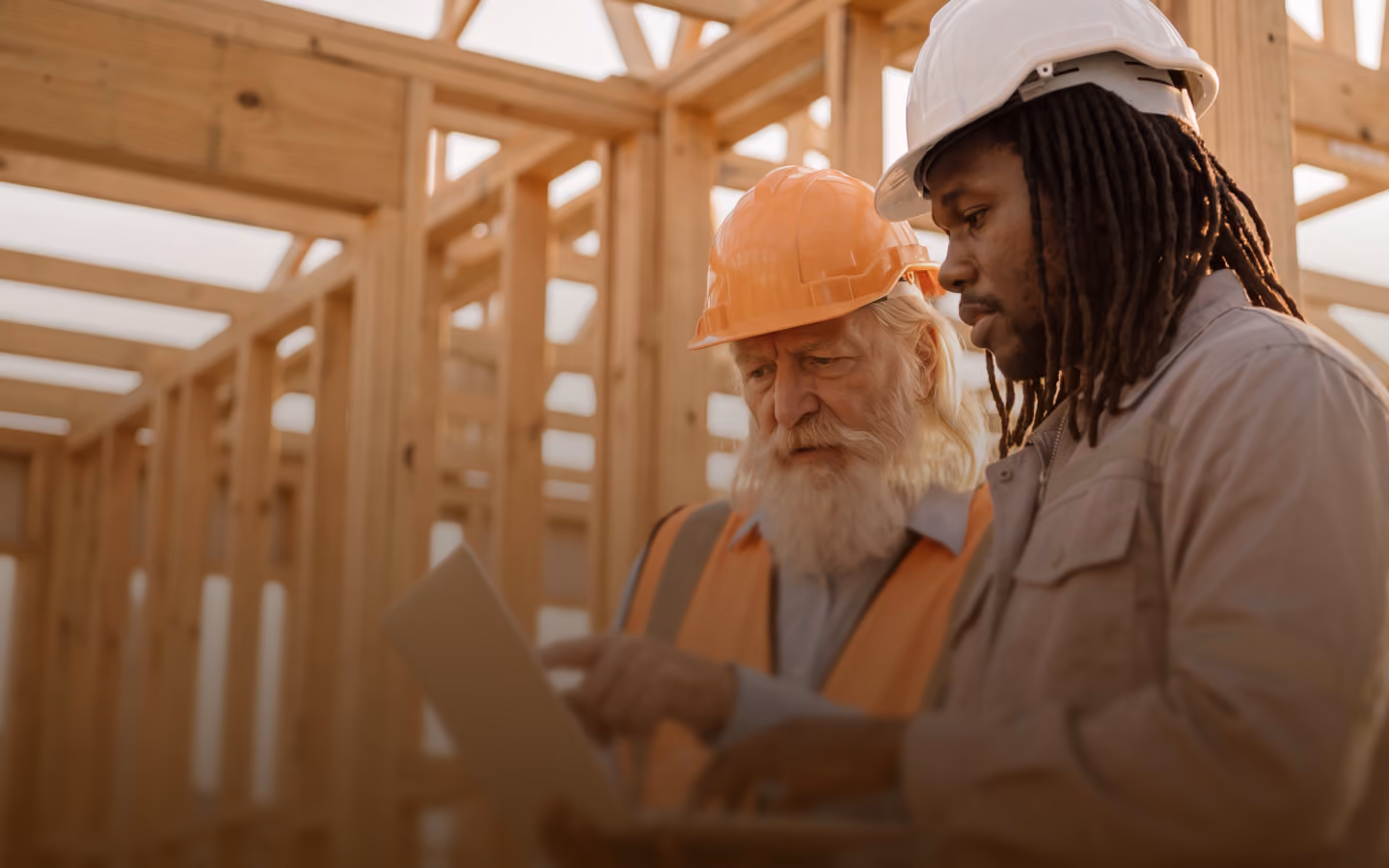 Two construction workers wearing safety helmets and work attire reviewing a document together inside a building framework made of wooden beams.