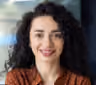 Smiling woman with curly dark hair wearing a brown and orange patterned top, posing indoors.