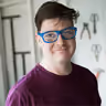 Person with short dark hair, wearing blue glasses and a dark maroon shirt, smiling indoors with blurred background containing tools on a wall.