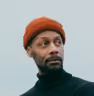 Portrait of a bearded man wearing an orange knit cap and a black turtleneck sweater looking slightly to the right against a plain gray background.
