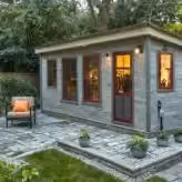 Small garden shed with three windows and a glass door, surrounded by a stone patio and green lawn, with an outdoor chair on the left.