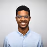 Smiling young Black man wearing glasses and a light blue button-up shirt, posing against a plain light gray background.