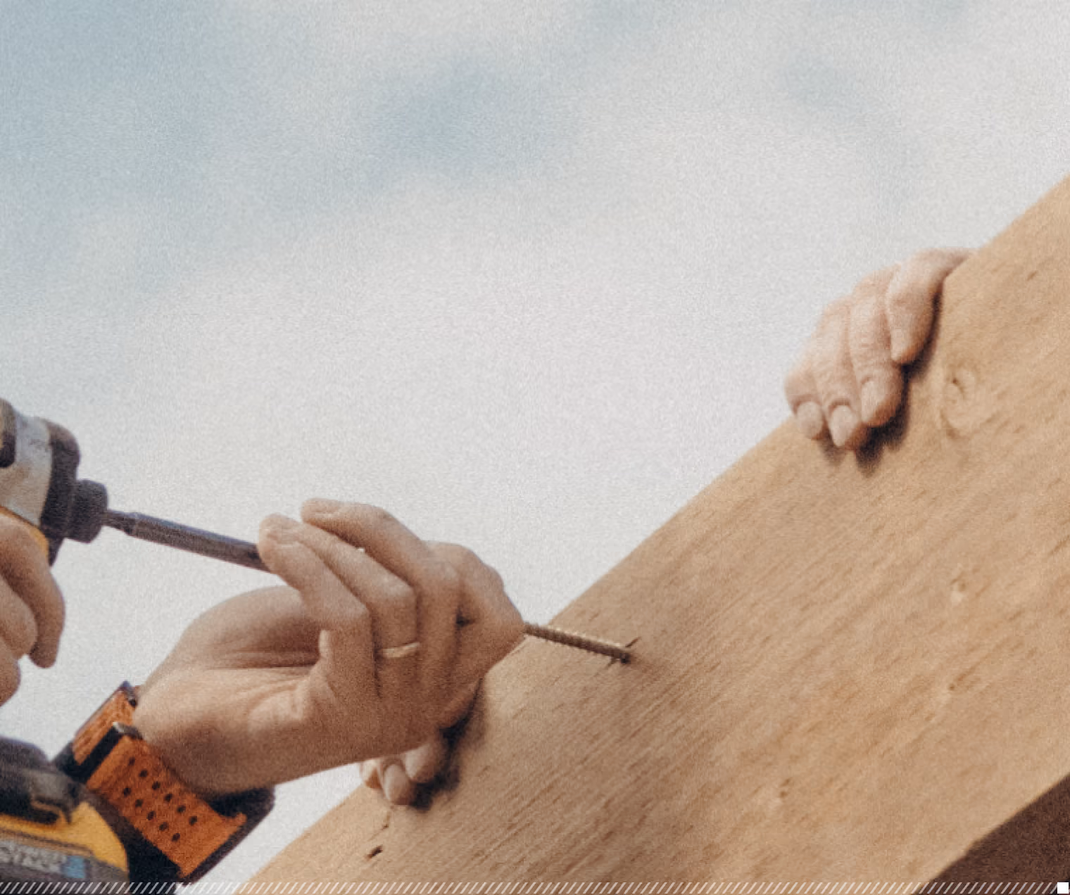 Hands using a power drill to screw a long screw into a wooden plank while another pair of hands hold the plank steady.