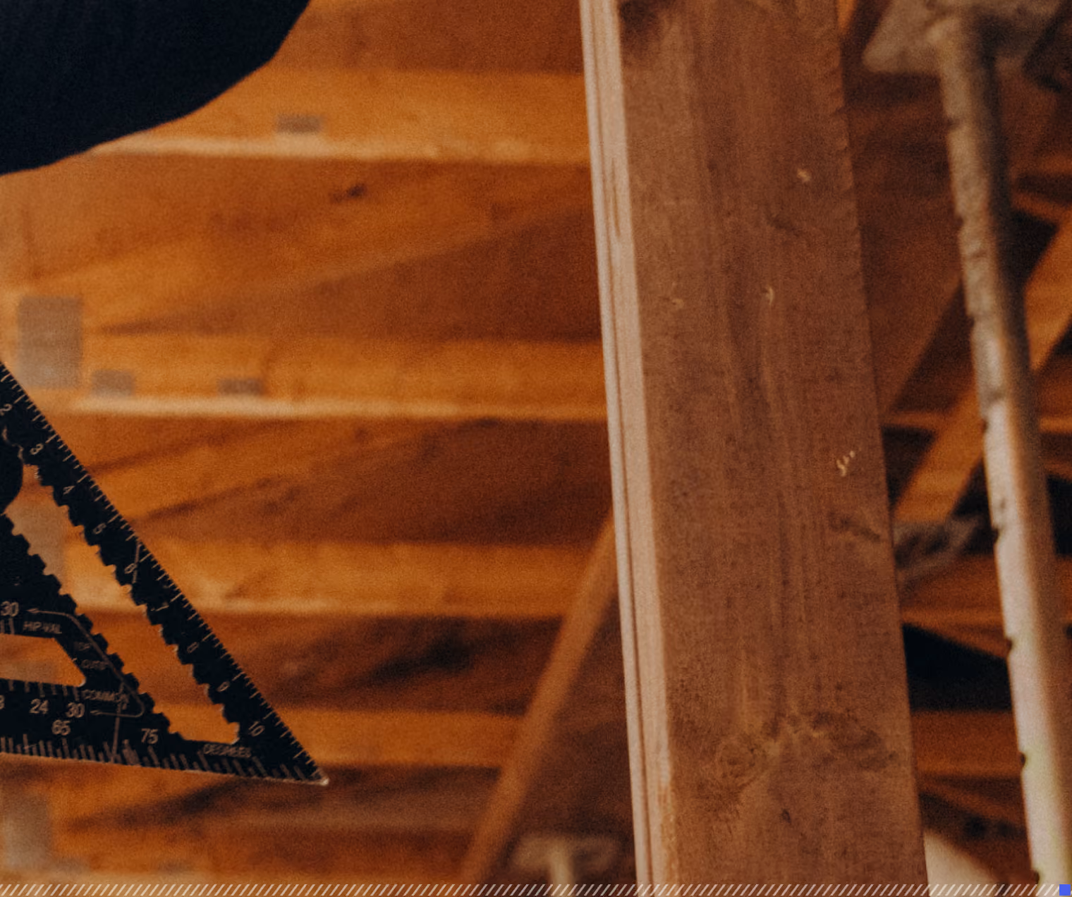 Person using a black carpenter's square to measure or mark a wooden beam in a construction setting with wooden rafters and support structures visible in the background.