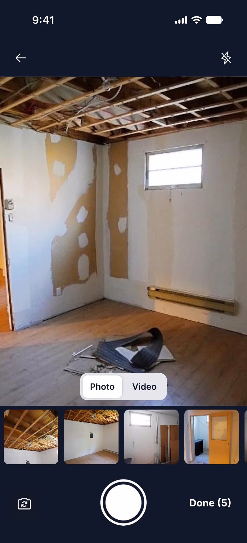 Photo of an unfinished room with exposed ceiling beams, partially patched walls, a small window, a wall heater, and debris on the wooden floor, displayed on a smartphone camera app interface from a photo gallery.