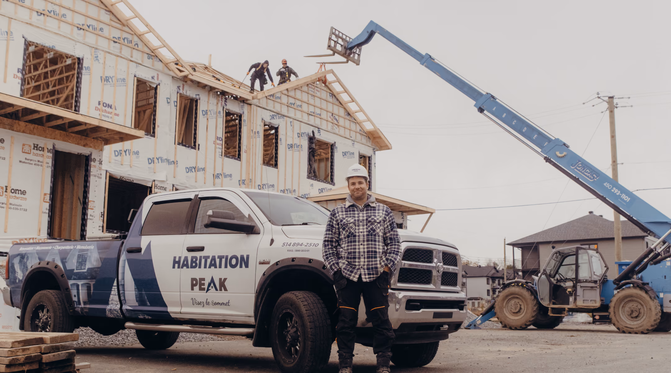 Construction site with a partially built house wrapped in protective sheeting, a white and blue truck labeled 'Habitation Peak' parked in front, a man wearing a hard hat and work clothes standing next to the truck, two workers on the roof, and a blue forklift lifting wooden materials.