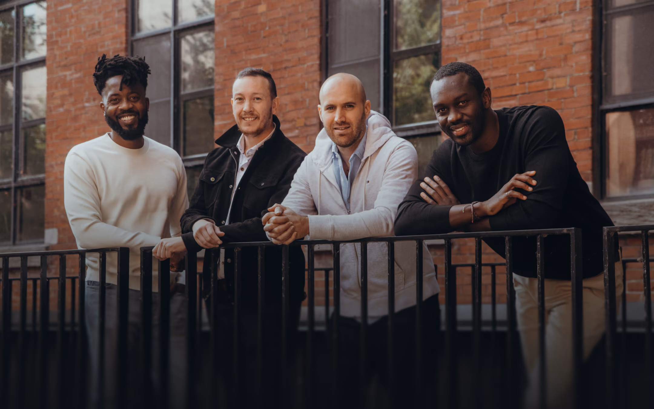 Four men leaning on a black metal railing in front of a brick building with windows, smiling and looking at the camera.