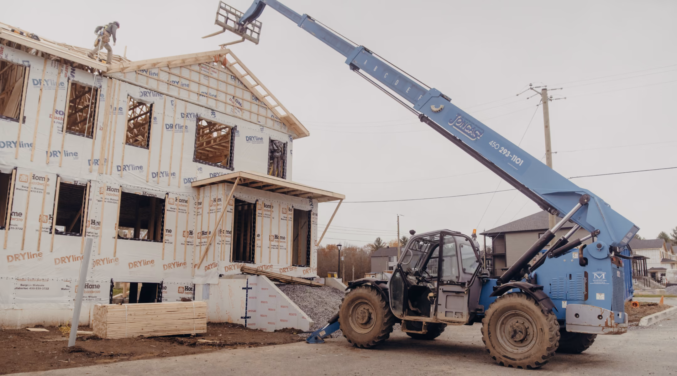 Construction site with a two-story house framed and covered in protective building wrap, a worker on the roof, and a blue telescopic forklift lifting materials.