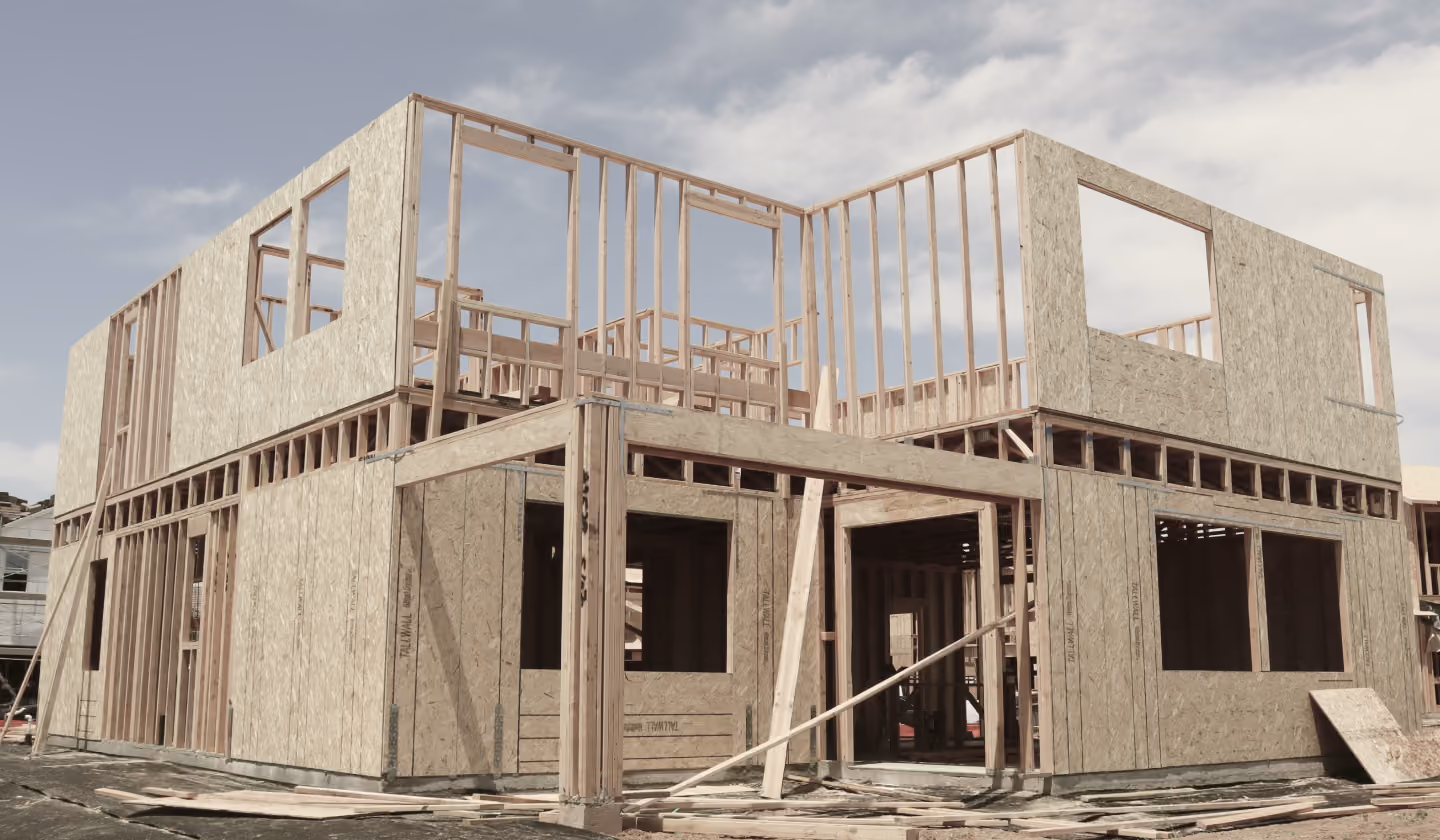 Two-story house under construction with wooden framework and OSB sheathing partially installed, showing window and door openings.