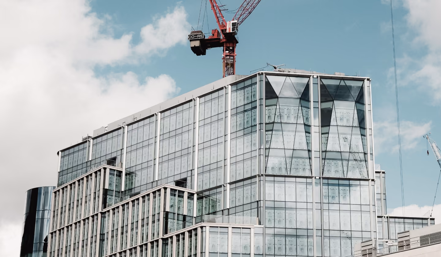 Modern glass office building with multiple levels and a red construction crane behind it under a cloudy blue sky.