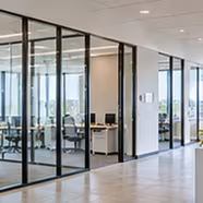 Modern office interior with glass-walled meeting rooms, white desks, office chairs, and computers inside, bright and spacious with tiled floors.