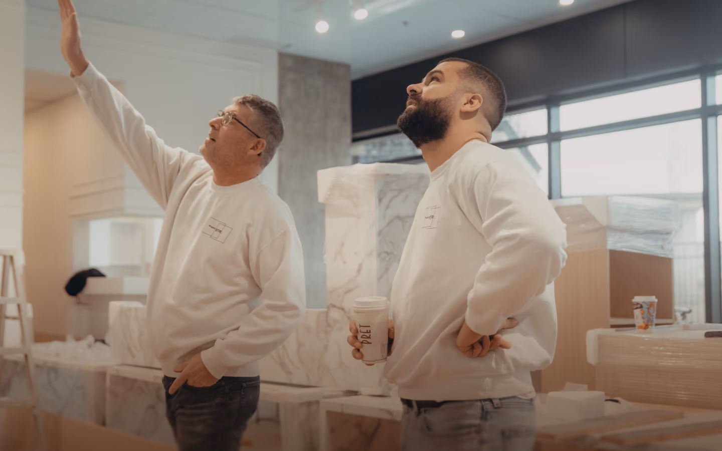 Two men wearing white sweatshirts inspecting a ceiling in a room filled with wrapped furniture and boxes, one man pointing upwards and the other holding a cup looking thoughtfully.