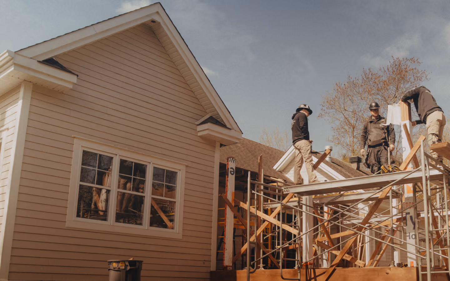 Three construction workers on scaffolding working on the exterior of a partially built beige house with white trim under a partly cloudy sky.