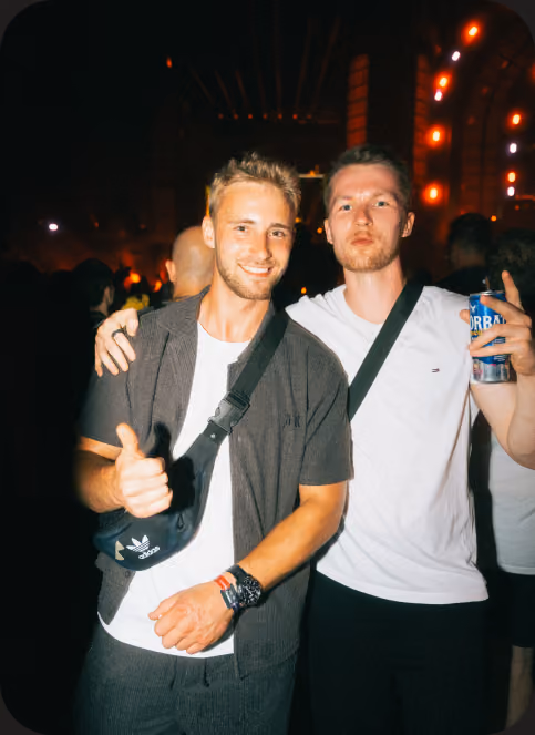 Zwei Männer bei Nacht auf einer Party, einer mit Daumen hoch, der andere hält ein Getränk, leuchtende Lichter im Hintergrund.