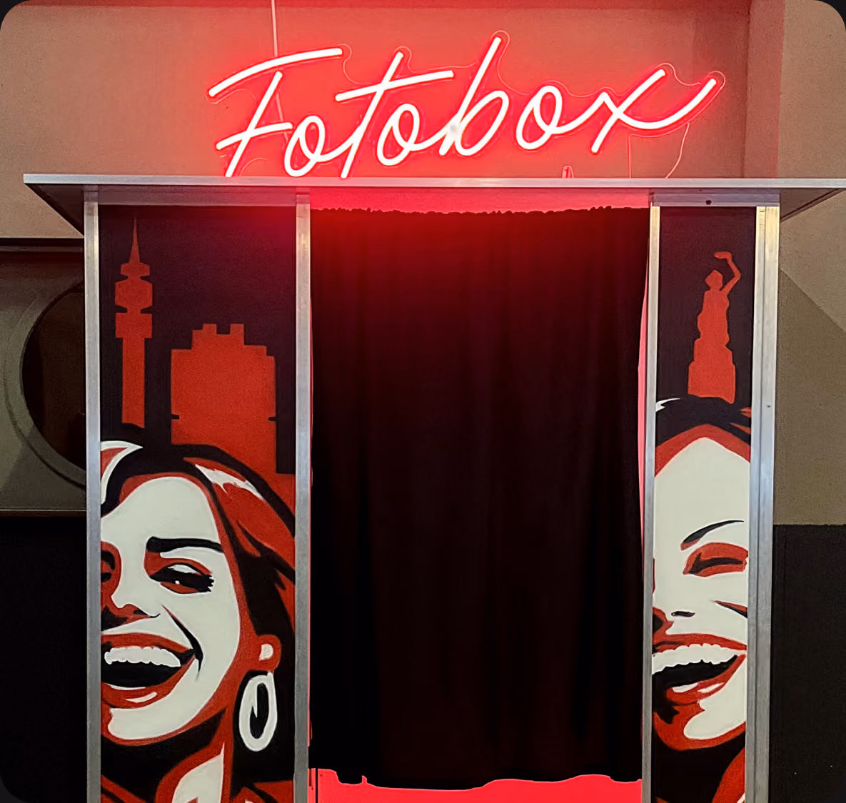 Rote Fotobox mit beleuchtetem Schriftzug, Vorhang und stilisierten lachenden Gesichtern auf rotem Hintergrund.