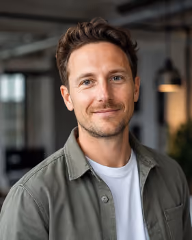 Mann mit braunen Haaren lächelt in einem modernen Büro mit hängender Lampe im Hintergrund.