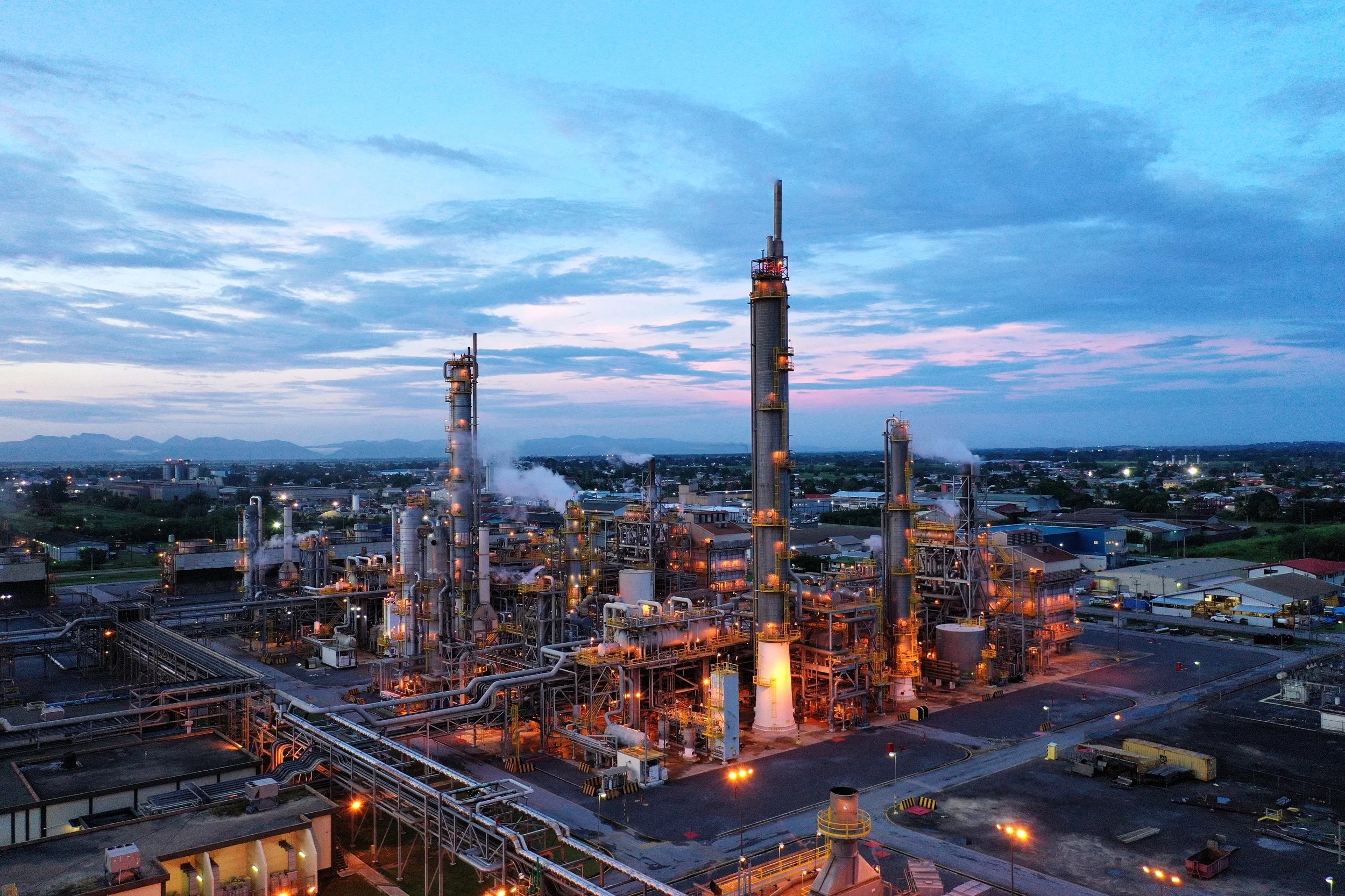 Aerial view at dusk of the Point Lisas industrial estate in Trinidad, its refinery towers and flare stacks lit against a blue and pink sky