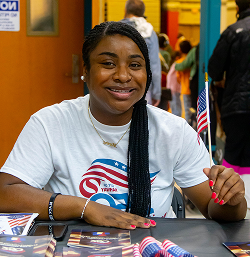 Smiling woman sitting at a table holding a small American flag in a busy indoor setting.
