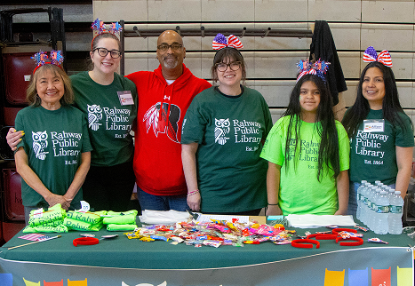 Six people at a Rahway Public Library table with books, candy, and water bottles, wearing festive headbands.