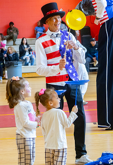 Man in patriotic outfit entertaining two children who watch a spinning yellow plate on a stick.