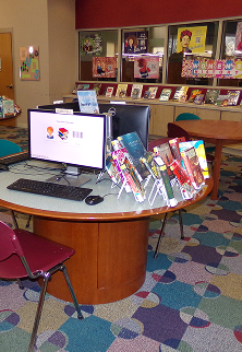 Computer on curved desk with colorful books displayed in a library with patterned carpet.