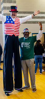 Man in tall stilts wearing red, white, and blue outfit with a hat stands next to a man in a green shirt.