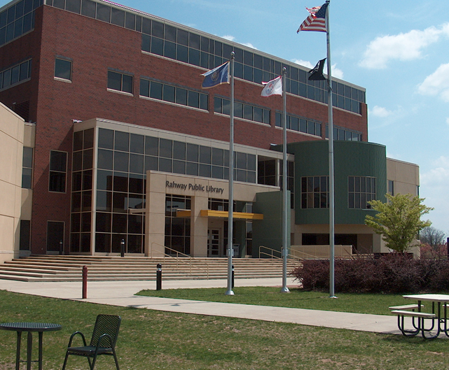 Modern Rahway Public Library building with flagpoles, lawn, benches, and chairs outside on a sunny day.
