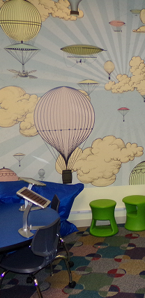 Room with colorful hot air balloon wall art, green stools, blue desk, and a chair with a tablet stand.