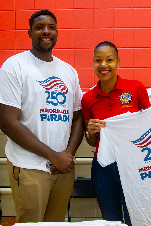 Smiling man and woman hold event T-shirts against a red tiled wall background.