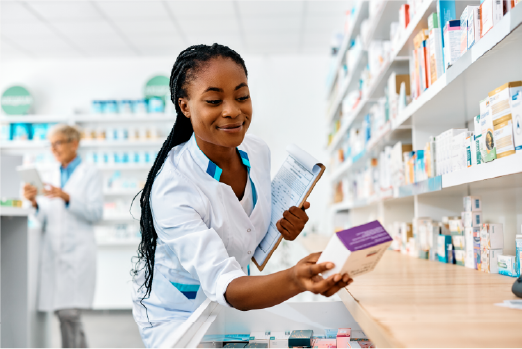 Female Black pharmacist holding a clipboard and selecting medication from pharmacy shelves.